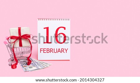 16th day of february. A gift box in a shopping trolley, dollars and a calendar with the date of 16 february on a pink background. Winter month, day of the year concept.