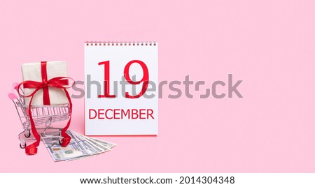19th day of december. A gift box in a shopping trolley, dollars and a calendar with the date of 19 december on a pink background. Winter month, day of the year concept.