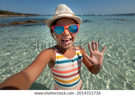 A happy carefree little girl is having fun to make a selfie or technology video call to parents or friends on a beach with seaside in a sunny day during family vacation holidays.