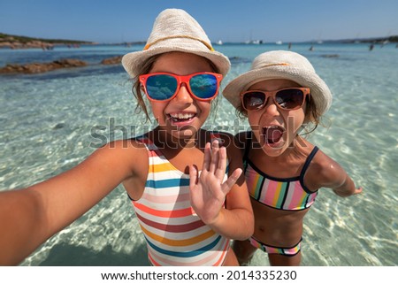 Happy carefree little girls sisters in bikini, straw hats and sunglasses making selfie or technology video call to friends or mother on seaside during family vacation holidays trip.Concept of roaming.