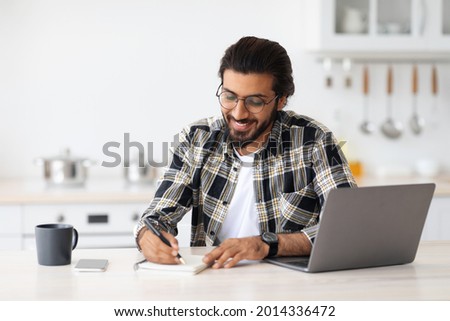 Positive arab guy with glasses looking for job online, sitting at table, using laptop and taking notes, kitchen interior, copy space. Excited indian man student working online from home, remote job