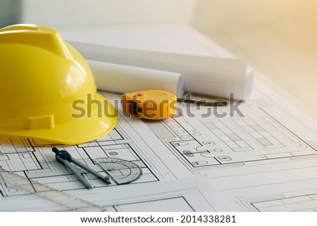 Yellow hard safety helmet hat and the blueprint, pen, ruler, protractor, and tape measure on the table at the construction site.for safety project of workman as engineer or worker, on city
