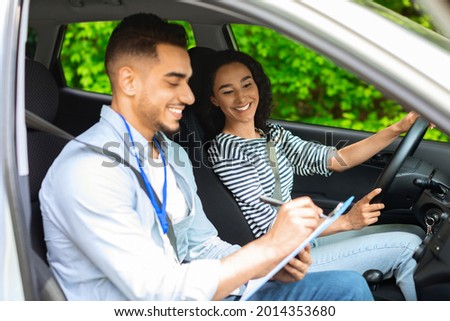 Cheerful middle-eastern guy instructor examinating happy brunette lady student, taking notes at chart while sitting by young woman driving car and waiting for test results, side view, copy space