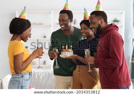 Cute millennial black friends making surprise for birthday woman, holding cake with lit candles, wearing party hats and smiling, greeting emotional african american lady, side view, home interior