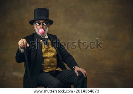 At cinema. Half-length retro portrait of elderly gray-haired man, gentleman or actor sitting on chair isolated on dark vintage background. Retro style, comparison of eras, humor concept. Looks