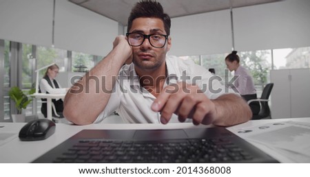 Bored or tired young bearded entrepreneur reading report or article on laptop screen. Portrait of indian male manager staring at computer screen feeling exhausted in office