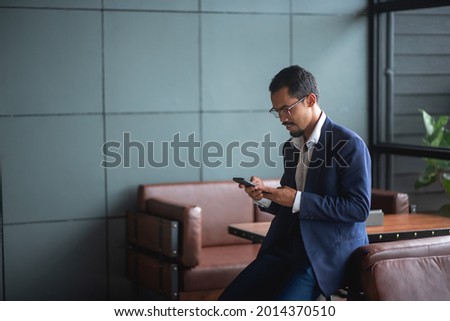 Young handsome businessman wears glasses and works on touchpad while sitting on sofa in office.