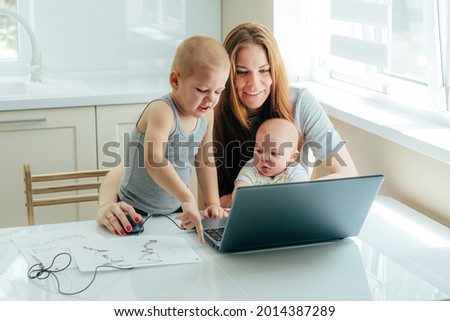 Young attractive mother with two children study online on a laptop in the kitchen at home.