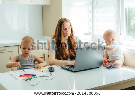 Young focused business mom with two children working remotely on a laptop at home in the kitchen.