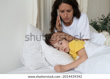 Worried mom calling to family doctor checking temperature of sick kid lying in bed. Small preschool boy suffer of fever from coronavirus or influenza sleep covered with blanket. Child cure and health Royalty-Free Stock Photo #2014439342