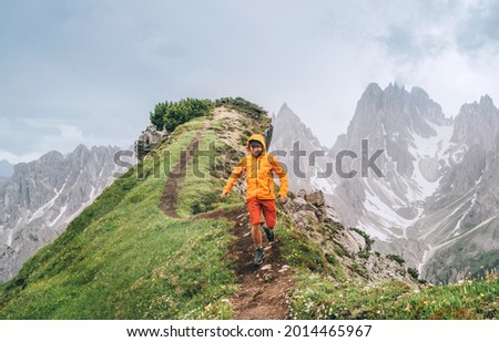 Dressed bright orange soft shell jacket backpacker running  by green mountain path with picturesque Dolomite Alps range background,. Active people and European mountain hiking tourism concept image. Royalty-Free Stock Photo #2014465967