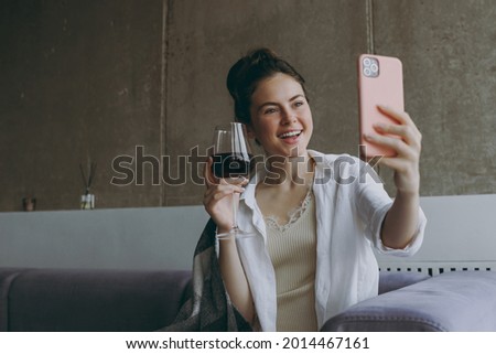 Young fun woman in white clothes plaid sitting on soft grey sofa indoors apartment hold glass drink red wine talk by mobile cell phone video call Rest on weekends leisure quarantine stay home concept