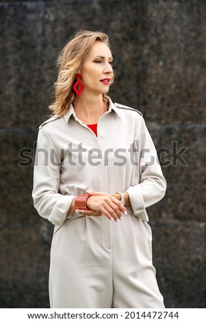 A woman in bright red diamond-shaped earrings and a beige elegant business jumpsuit, a wide red bracelet on her arm. Business-style clothing for everyday life. Royalty-Free Stock Photo #2014472744