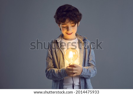 Little smiling curly boy stands on a gray background and looks at a bright light bulb in his hands. Concept of creative thinking and early development of intelligence. Banner. Place for text.
