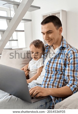 Happy young adult dad embracing cute small kid son looking at laptop device sitting on couch. Smiling father teaching toddler child using computer at home learning online, playing, watching cartoons.