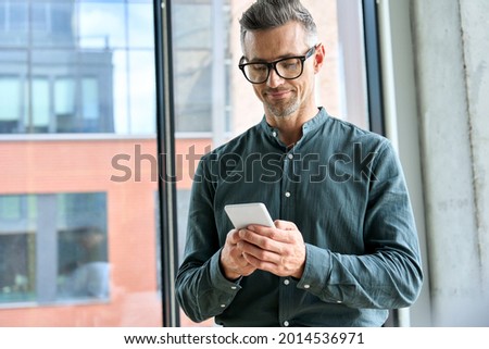 Smiling mature businessman holding smartphone standing in office. Middle aged manager ceo using cell phone mobile apps. Digital technology applications and solutions for business corporate development Royalty-Free Stock Photo #2014536971