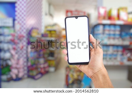 Hand holding smartphone with blank white screen, blurry supermarket view in the background, online shopping concept, mockup image.
