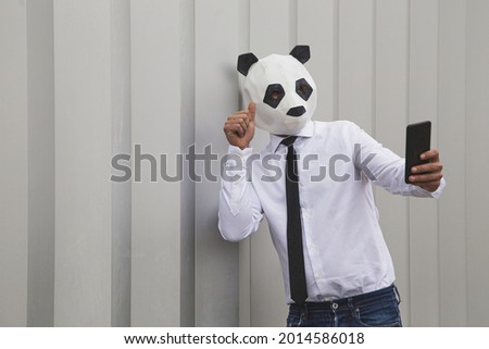 A male in a white shirt, tie, and panda head mask taking smartphone selfie against the gray wall