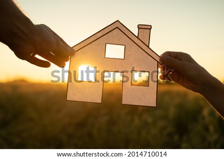 Family holds paper house at sunset, sun shines through window. Hand holding paper cut of house symbols at sunset 