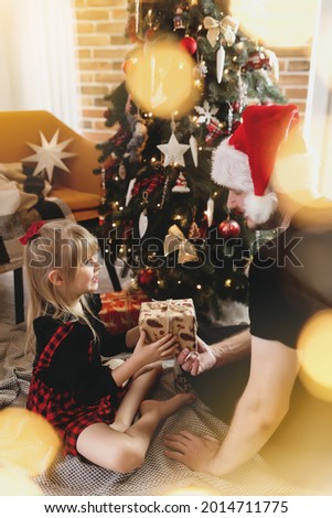 Young father in red Santa hat giving Christmas gift to his smiling daughter near decorated Christmas tree. Girl dressed in festive red-black Christmas outfit. They smiling, happy because of New Year