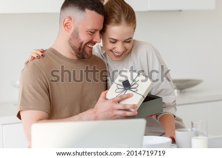 Young loving wife making surprise for husband for anniversary or birthday, congratulating him and giving present while having healthy breakfast in morning at kitchen, happy man opening gift box Royalty-Free Stock Photo #2014719719