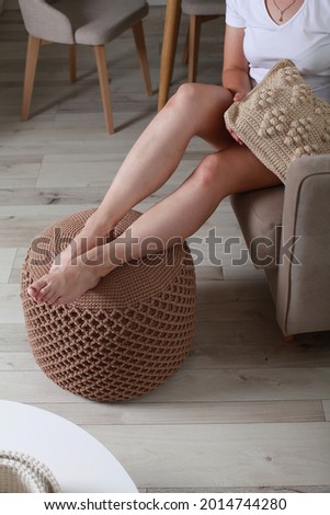 Cropped close up image of female legs resting on Stylish handmade knitted pouf in interior. Natural jute round pouf cushion chair with wooden legs, grey floor. Close up details. Minimalist 