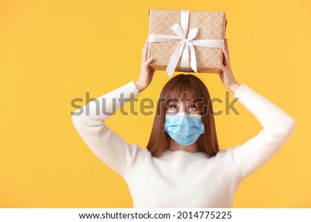 Beautiful young woman in medical mask and with gift on color background