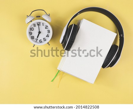 Top view or flat lay of white books  with headphones around and white vintage alarm clock shows 7 o'clock on yellow background. Audio books, morning routine concept.