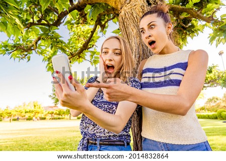Two girls making surprised faces with mouths and eyes wide open pointing and looking smartphone spending time in city park nature. Young women having fun with social networks due to wi-fi technology