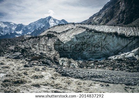 Scenic landscape with powerful mountain river beginning from glacier among large moraines on background of great snowy mountains. Beautiful scenery with glacier at source of turbulent mountain river.