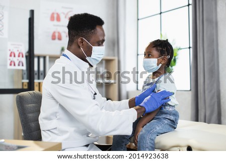 Competent male pediatrician in medical mask and gloves making Covid-19 vaccination in shoulder of little african girl. Concept of immunisation, protection and medicine. Royalty-Free Stock Photo #2014923638