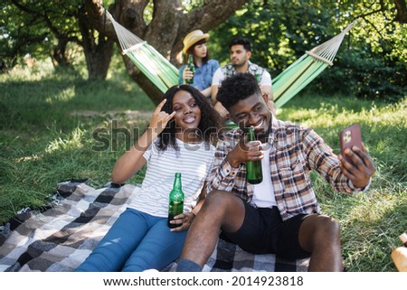 Happy african couple sitting together on plaid at park and taking selfie on cell phone while international friends drinking beer in hammock on background. Relaxation of young people.