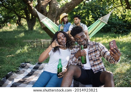 Funny black man amd woman relaxing at summer park and taking selfie on mobile. Blur background of two diverse friends sitting in hammock with beer in hands.