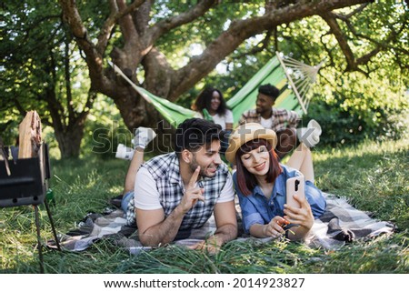 Cheerful couple taking selfie on smartphone during picnic outdoors. Blur background of african man and woman sitting in hammock and drinking beer. Summertime and friendship concept.