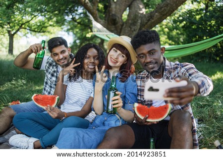 Cheerful group of four diverse friends sitting at green garden with watermelon and beer in hands and taking selfie on modern smartphone, Concept of people, technology and leisure time.