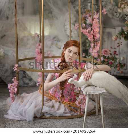 Red-haired girl sits in a golden cage with a white peacock on a stool