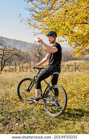 Active man riding bicycle at autumn day. Male bicyclist holding cellphone and taking picture of beautiful autumn hills. Concept of autumn sport travel, mountain biking, active leisure