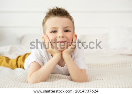 Happy Child boy in a white T-shirt smiles and looks into the camera while lying on the bed, supports his head with his hands. Kid Boy close-up on a white background, looking into the camera.