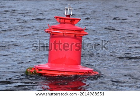 Red buoy on the undulating surface of the river. Lantern at the top of the buoy. The lower part is overgrown with green algae.