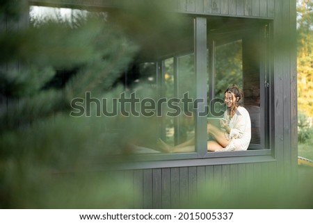 Young woman resting at beautiful country house or hotel, sitting with tablet on the window sill enjoying beautiful view on pine forest. View from outside. Beautiful destinations for vacation Royalty-Free Stock Photo #2015005337