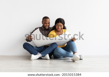Young African American Spouses With Blueprints In Hands Sitting On Floor In Their New Apartment, Excited Black Couple Looking At Building Plan And Thinking About Interior Design, Copy Space Royalty-Free Stock Photo #2015015384