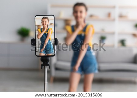 Young Blogger Concept. Cheerful teen girl dancing at camera filming video using phone on tripod at home, creating her trendy content on a mobile app to share on social media, selective focus on screen Royalty-Free Stock Photo #2015015606