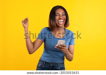 Happy Black Lady Holding Phone Gesturing Yes Winning Mobile Game Over Yellow Background. Studio Shot Of African Woman Celebrating Great News Posing With Cellphone Royalty-Free Stock Photo #2015015615