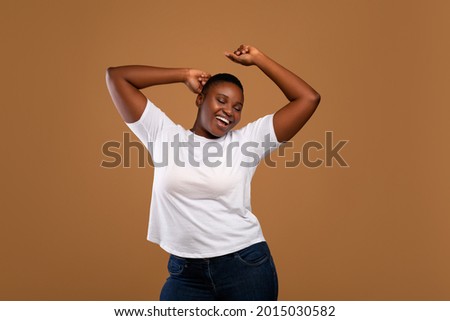 Taking Break. Overjoyed millennial plus-size black woman wearing white t-shirt dancing with closed eyes, having fun moving and listening to music, relaxing isolated over dark brown studio background Royalty-Free Stock Photo #2015030582