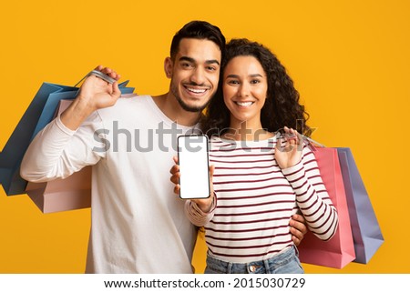 Online Shopping App. Cheerful young arab couple showing smartphone with blank white screen at camera and holding bright shopper bags while standing isolated over yellow studio background, mockup Royalty-Free Stock Photo #2015030729