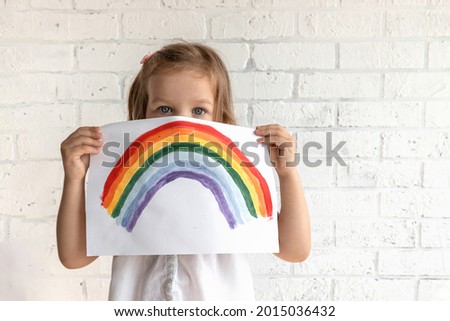 A little beautiful girl looks at the camera and holds a sheet of paper on which a rainbow is drawn. Pandemic hope concept. Stay at home