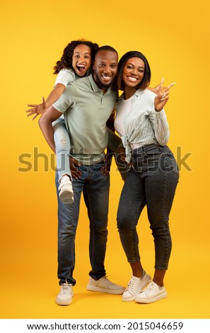 Joyful african american family with cute daughter posing over yellow background, excited parents having fun with their child, girl piggybacking dad and smiling. Full length portrait