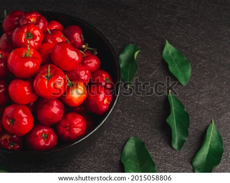 Top view of red acerola cherries fruit in a ceramic bowl with green leaves on a stone table. High vitamin C and antioxidant fruits. Close-up. Space for text. Concept of healthy fruits.