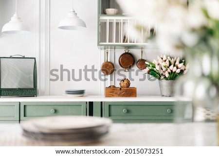 Set of plates and bouquet of flowers in glass jar on countertop in blurred foreground, green furniture with drawers and hanging pendant lights, white themed cozy kitchen Royalty-Free Stock Photo #2015073251