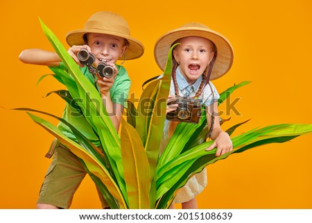 Young travelers, a girl and a boy, explore nature, they look out from behind palm leaves with binoculars and a camera. Summer vacation, tourism. Studio portrait on a yellow background.  Royalty-Free Stock Photo #2015108639